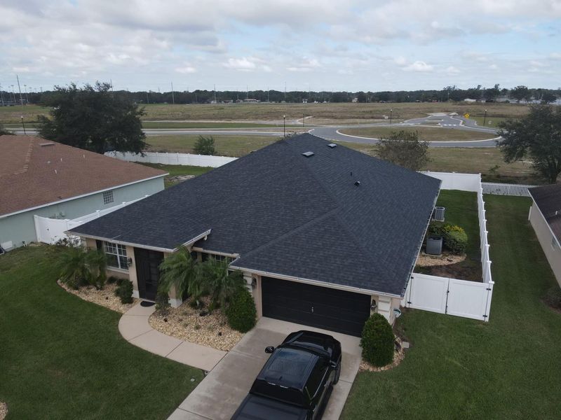 Aerial view of a house with a dark roof, black car in driveway, white fence, and green lawn.