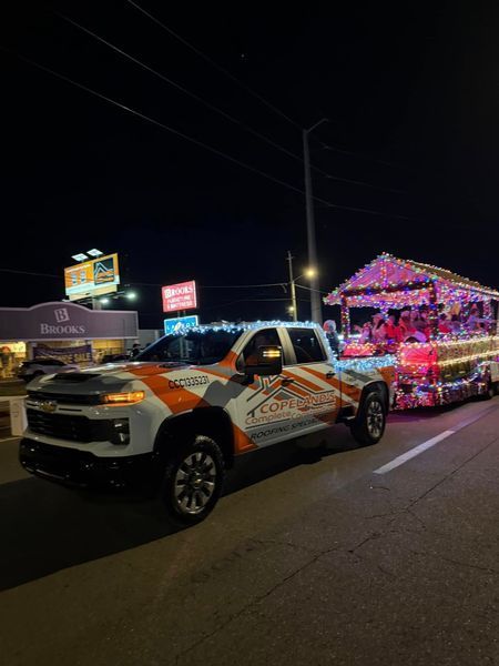 A white Chevrolet truck with an orange wrap pulls a trailer decorated with Christmas lights at night.