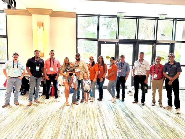 Group of people posing indoors in front of windows. Some hold children; many wear lanyards.