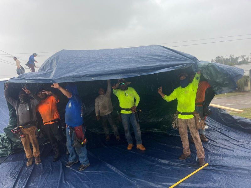 People on a roof holding up a blue tarp. Men wearing work clothes in overcast weather.
