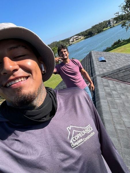 Two men smiling on a rooftop, one taking a selfie, overlooking a lake. Bright sunny day.