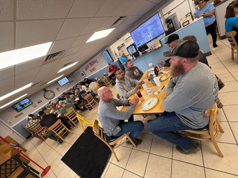 A crowded diner. Men seated around a table, some talking and smiling. Others eat. Blue walls, a menu, and yellow caution tape.
