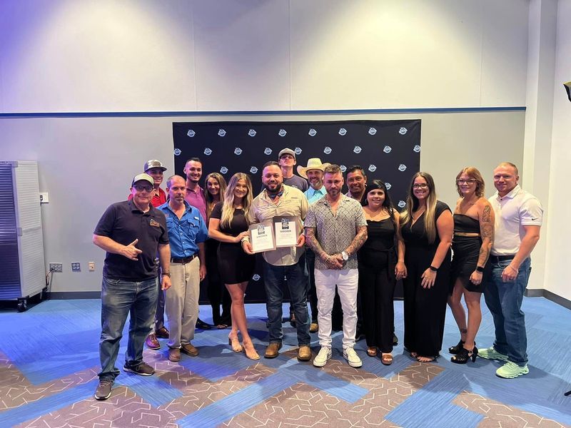 Group of people posing with awards in front of a blue backdrop, smiling and celebrating.