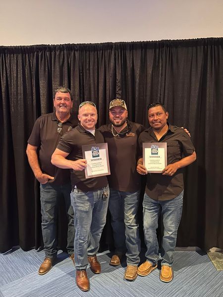Four men in brown shirts and jeans holding awards, smiling in front of a black curtain.