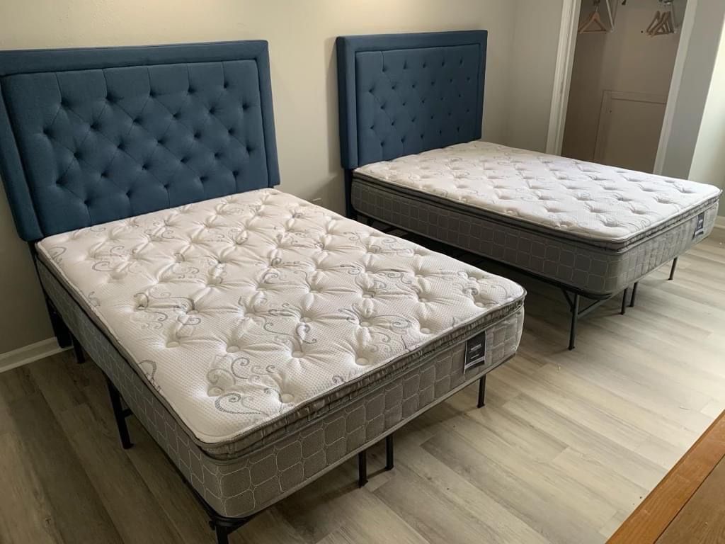 Two twin beds with tufted blue headboards and white mattresses in a bedroom with light-colored wood flooring.