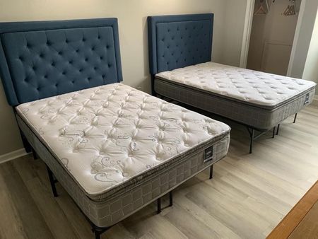 Two twin beds with tufted blue headboards and white mattresses in a bedroom with light-colored wood flooring.