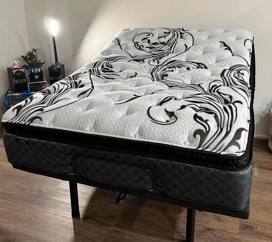 A white and black patterned mattress on a black metal frame; a lamp, plant, and electronics are to the left on the floor.