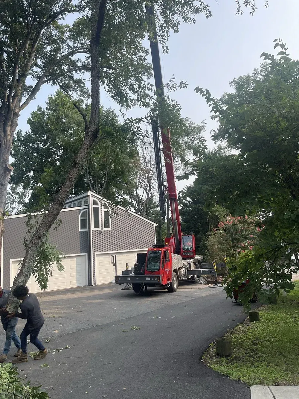 Red crane truck lifting debris on a tree-lined residential street beside a gray house