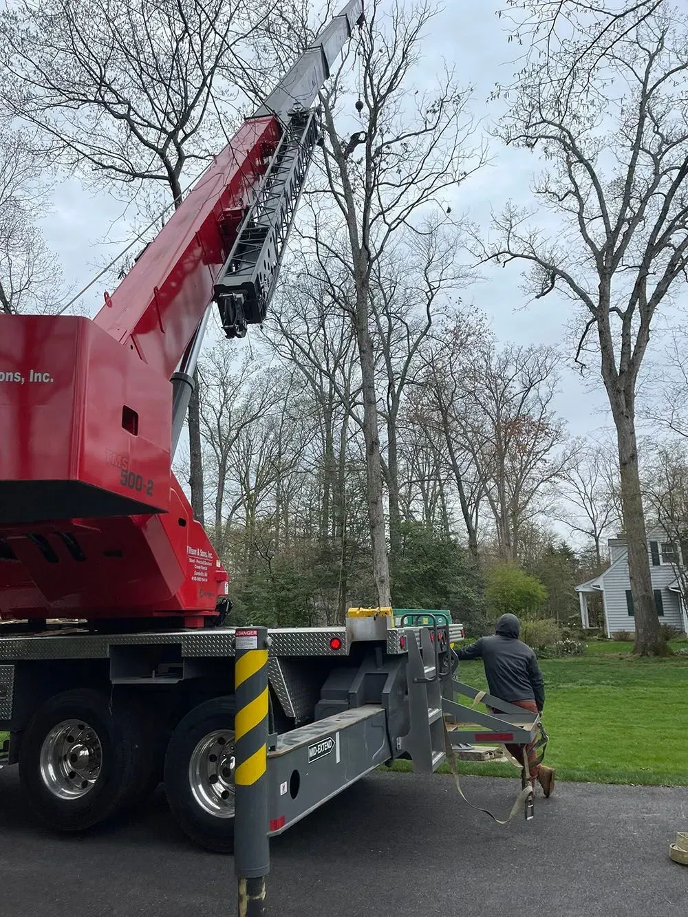 Red crane truck lifting debris by a roadside with a worker nearby in a wooded residential area