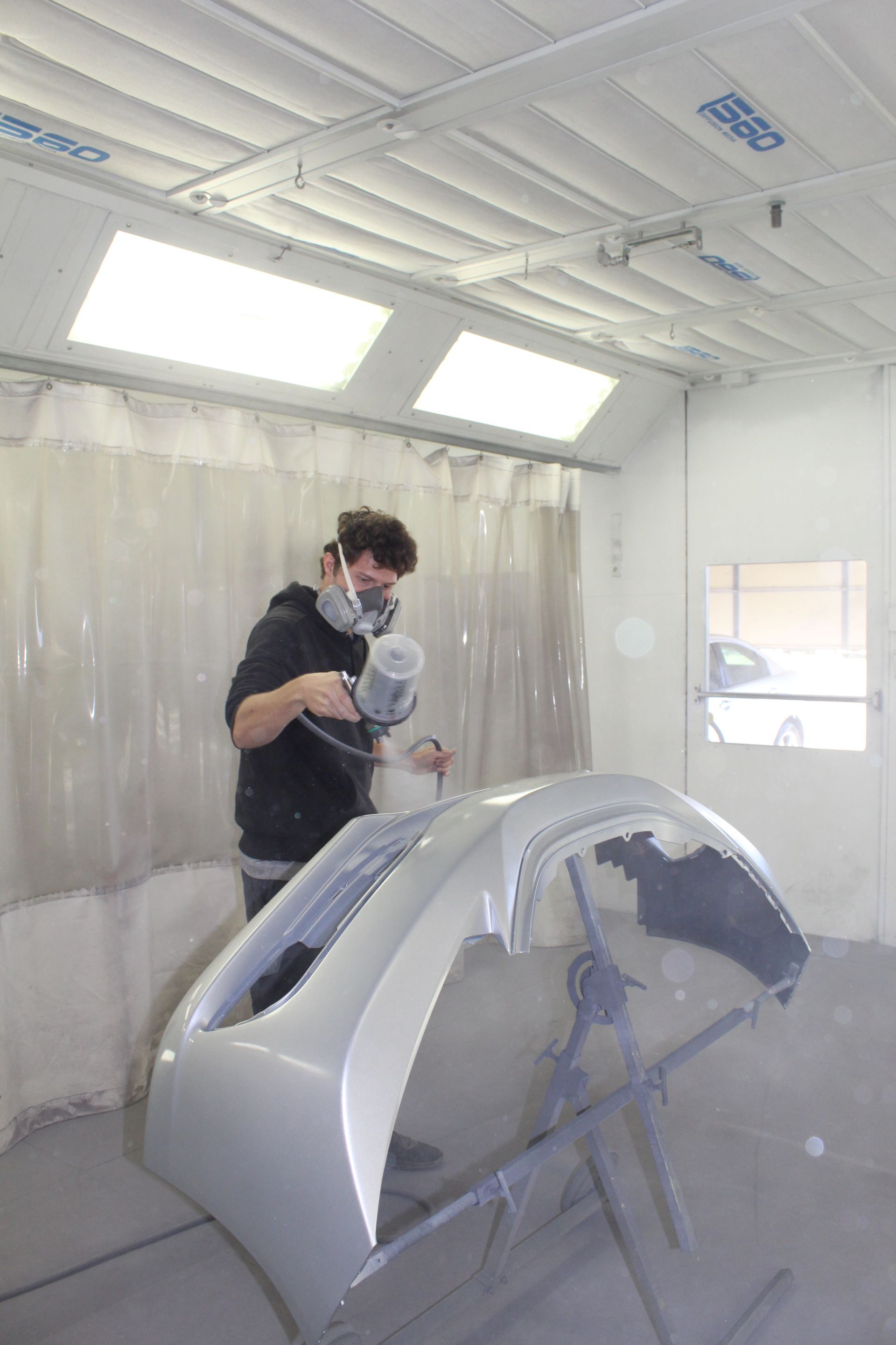 A man is spray painting a car bumper in a paint booth