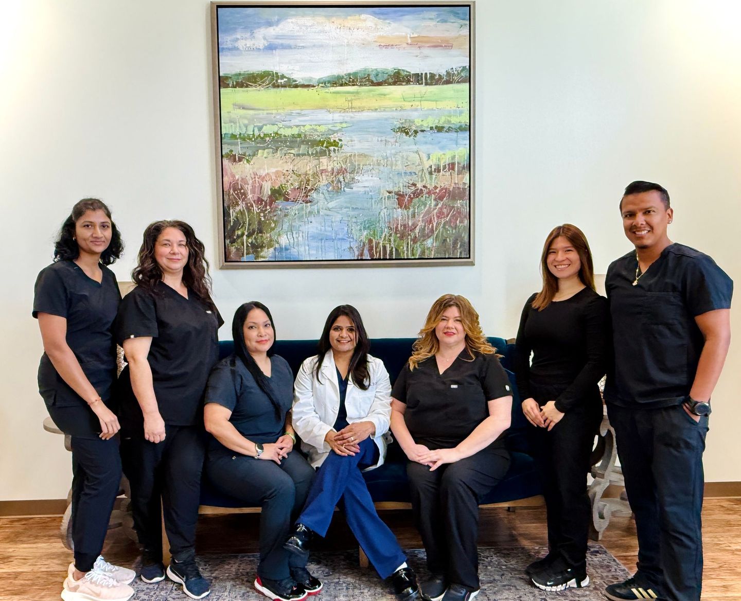 Medical staff in scrubs pose for a group photo in an office setting, with a painting on the wall.