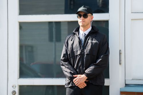 Man in black jacket and cap standing outside a white building with hands clasped
