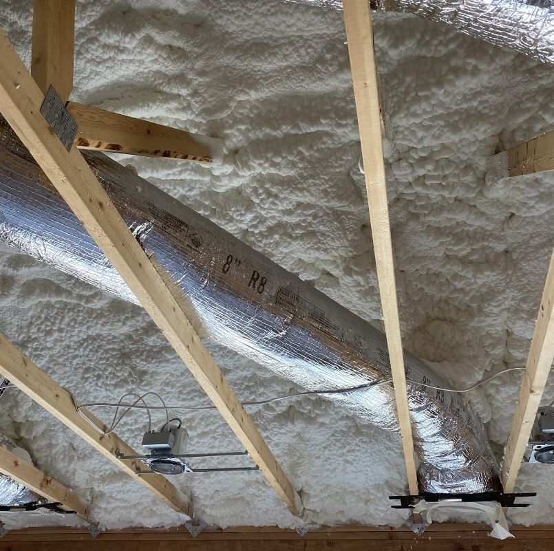 Spray foam insulation covers a wooden attic ceiling with a metallic duct.