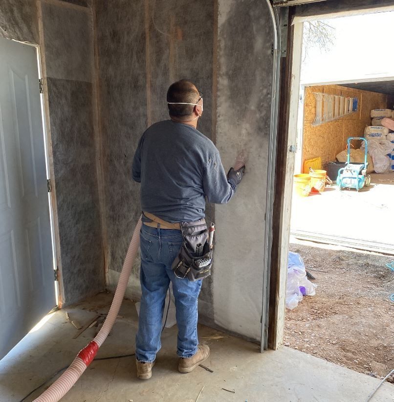 Man in gray shirt insulates a garage wall, using a hose connected to a machine.