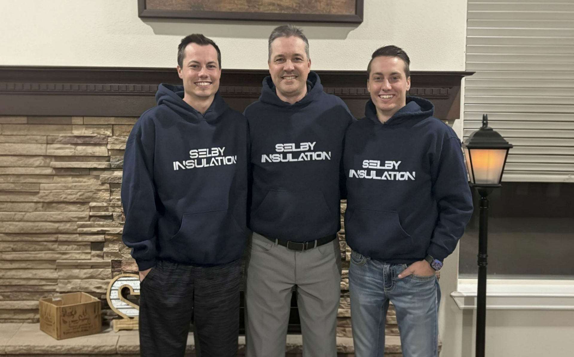 Person in protective suit spraying insulation into an attic. Wooden beams and rafters are visible.