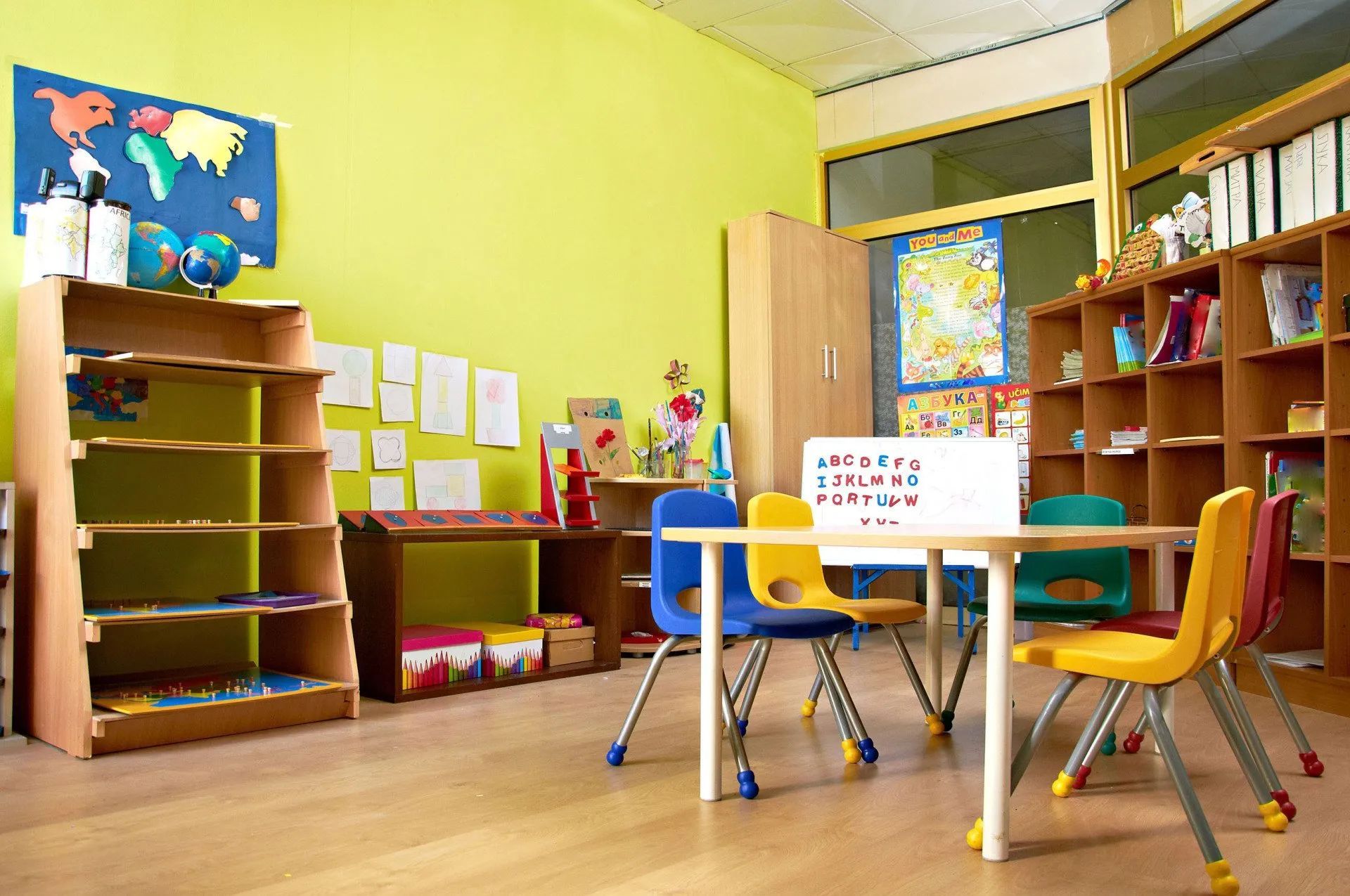 Bright preschool classroom with colorful chairs, tables, shelves, and children's artwork on yellow walls