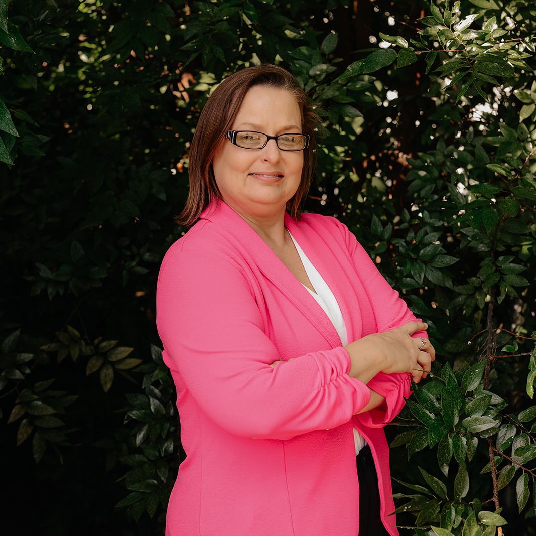 A smiling person wearing glasses and a bright pink blazer, standing with arms crossed in front of a leafy green background.