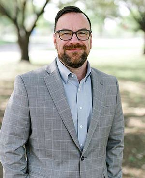 Bryan Campbell in a suit and glasses is standing in a park with his hands in his pockets.
