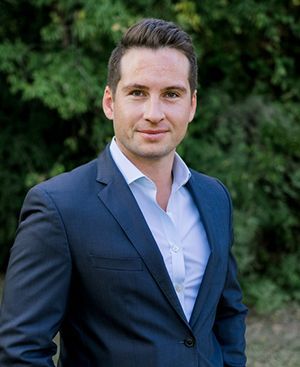 James Davidson in a suit and white shirt is standing in front of trees.