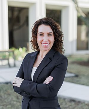 Lori Steenken in a black suit is standing in front of a building with her arms crossed and smiling.