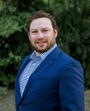 Matthew Davidson in a blue suit and plaid shirt is standing in front of trees.