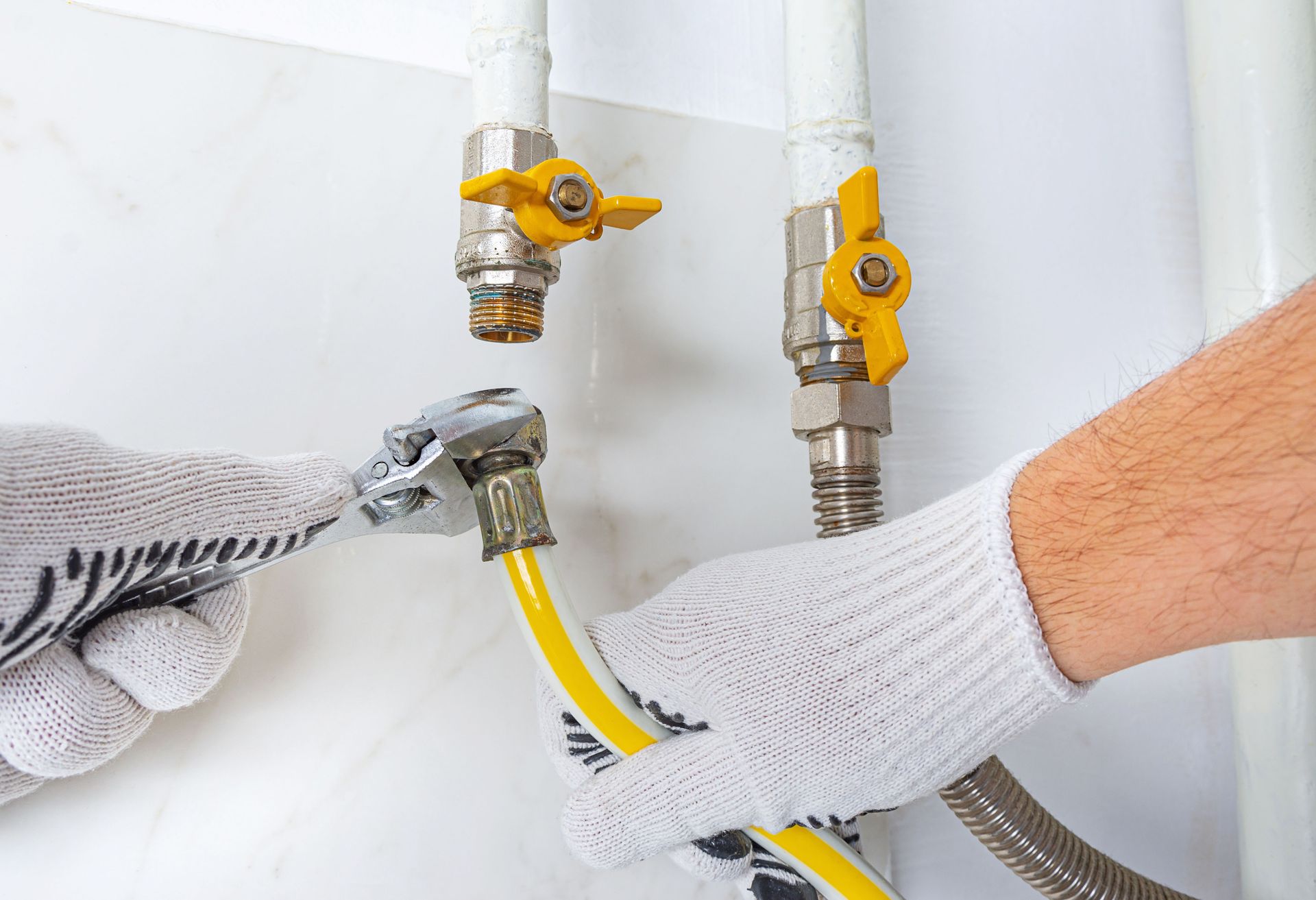Gloved hands connect a yellow gas hose to metal shutoff valves on a white wall.