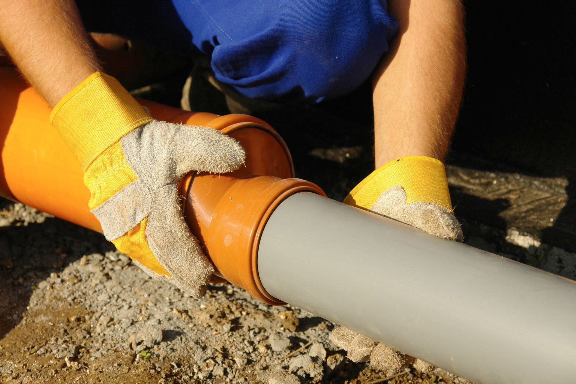 Gloved hands connecting an orange pipe fitting to a gray PVC pipe outdoors