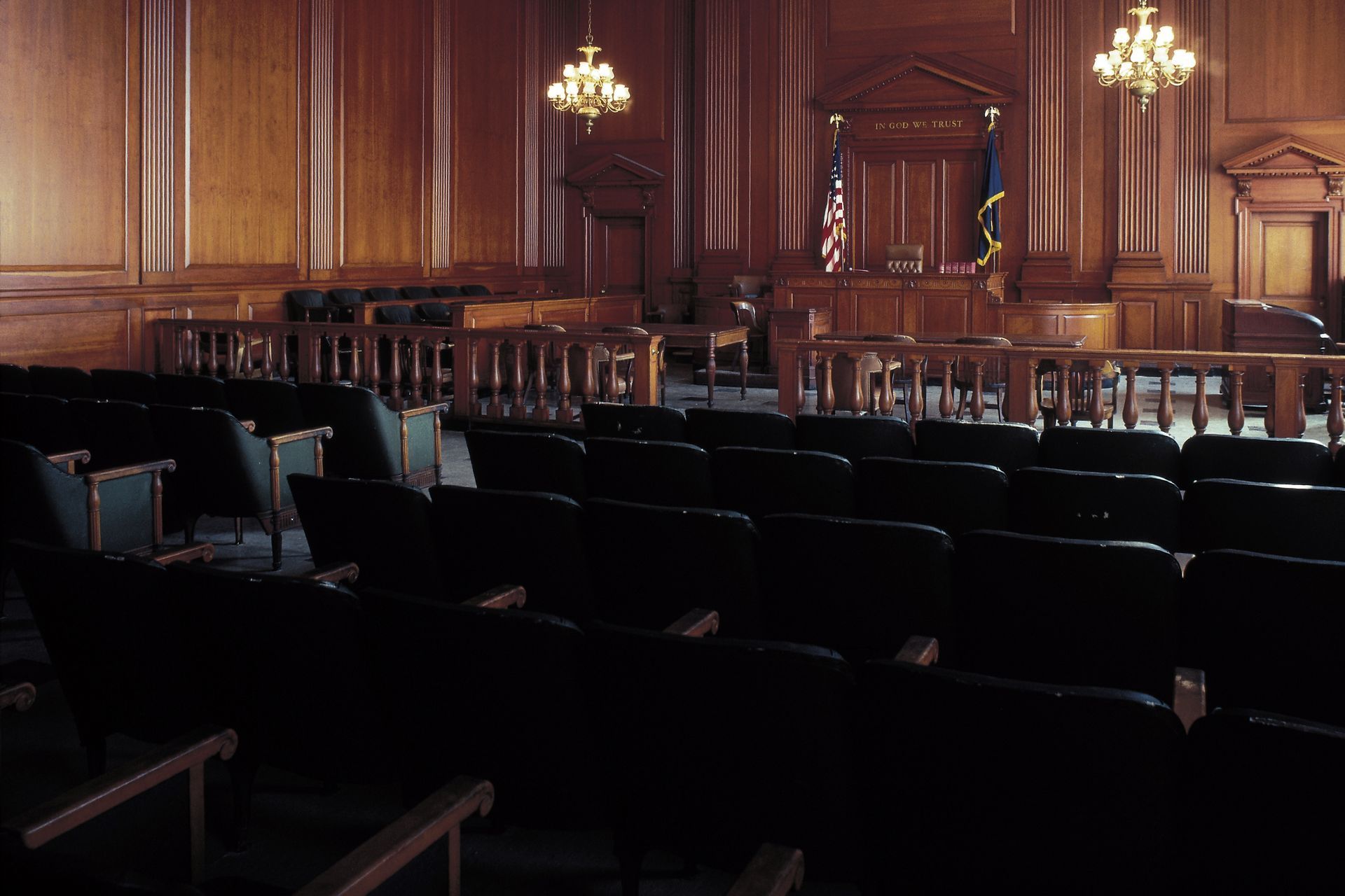 An empty courtroom with rows of dark chairs facing a judge's bench and wood-paneled walls under two chandeliers.