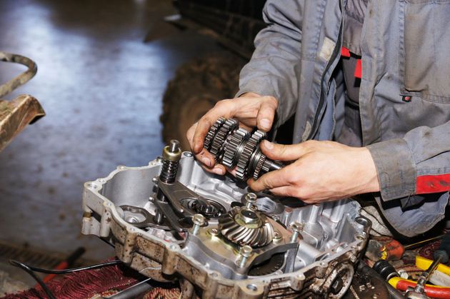 A mechanic wearing grey coveralls installs a gear assembly into an open vehicle transmission housing.