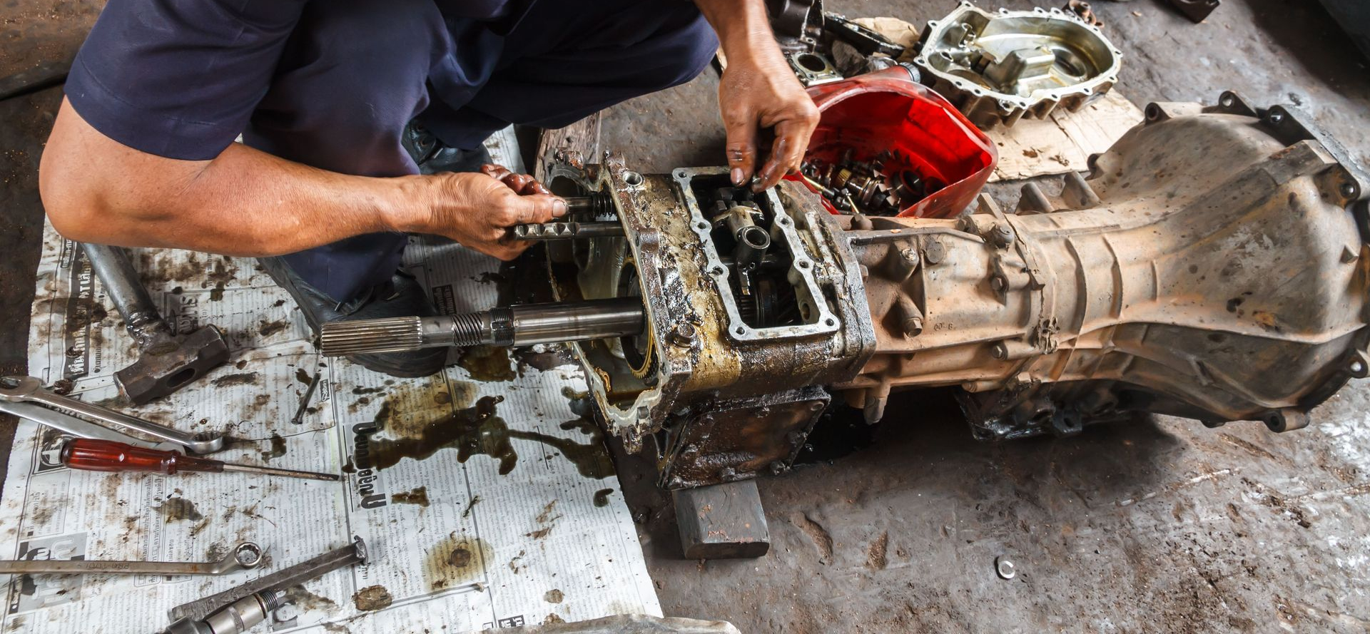 A mechanic repairs a dirty manual car transmission on the ground in a workshop.
