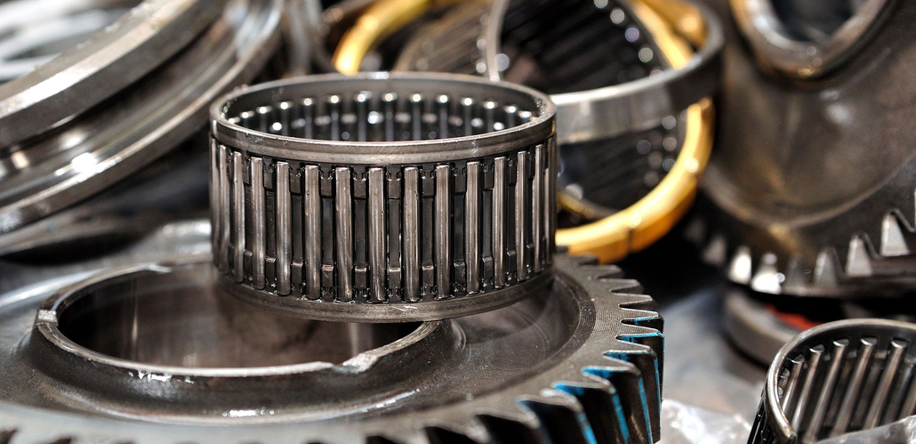 A close-up of a cylindrical needle bearing resting atop metal gears and mechanical parts in a workshop setting.
