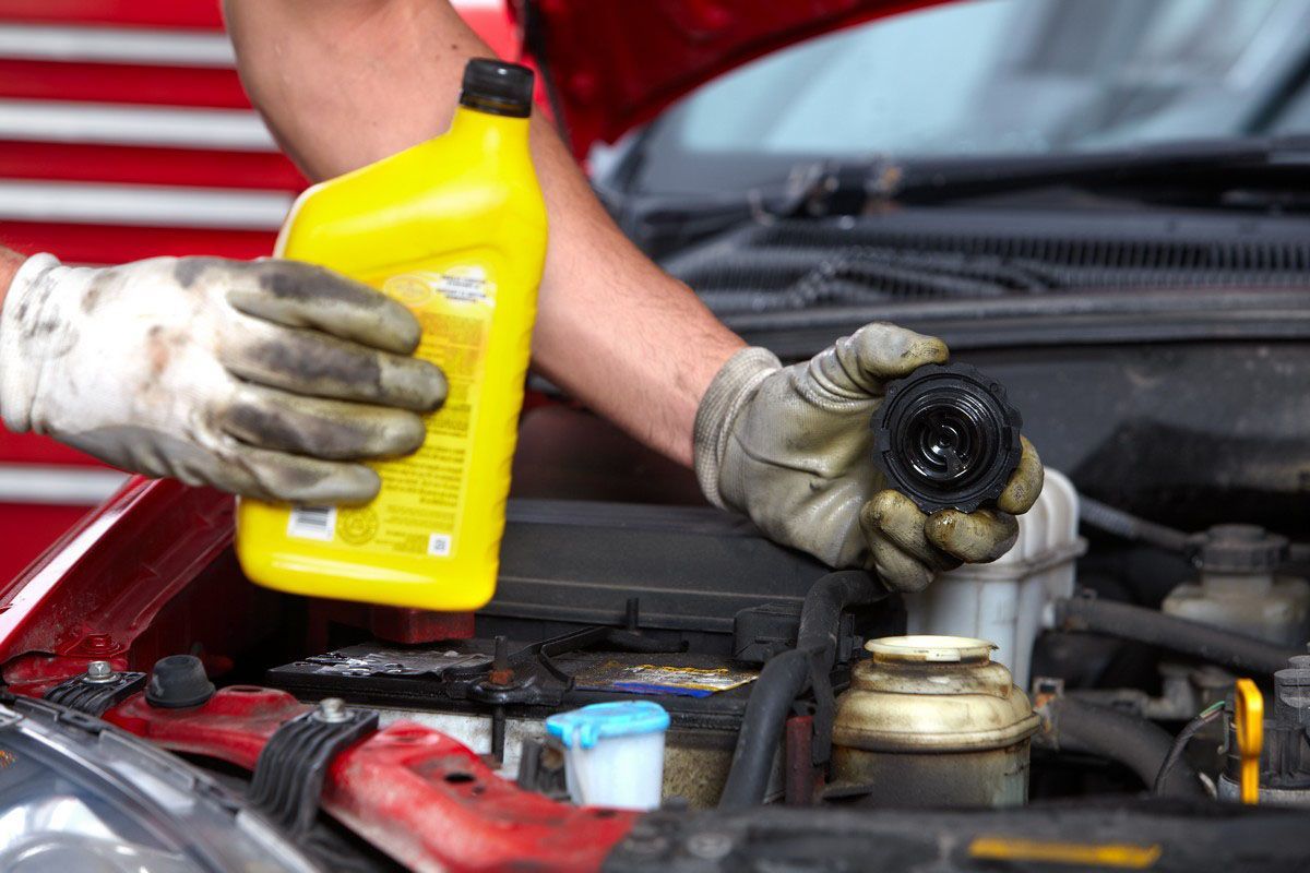 A pair of gloved hands holding a yellow motor oil bottle and a black engine oil cap over a car engine.