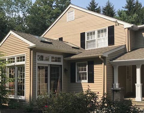 Tan house with dark shutters, multiple windows, and a covered porch on a sunny day.