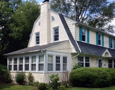 Beige two-story house with black roof, blue shutters, and a porch, set amongst green trees and bushes.