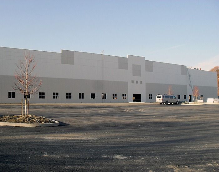 Large industrial building with a paved lot; two-tone gray facade, several loading docks, and a few parked vehicles.