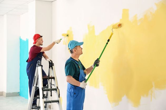 Two people painting a white wall yellow; one on a ladder, the other standing on the floor, both using paint rollers.
