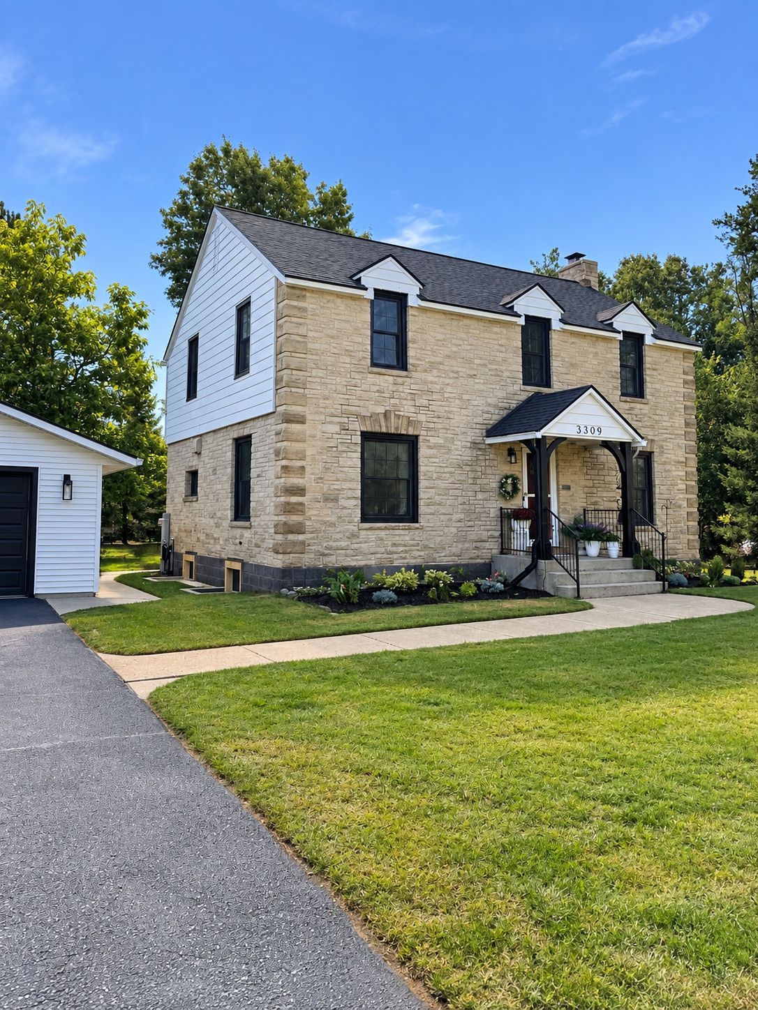 Two-story tan stone house with black shutters and a small front porch, beside a driveway and green lawn.