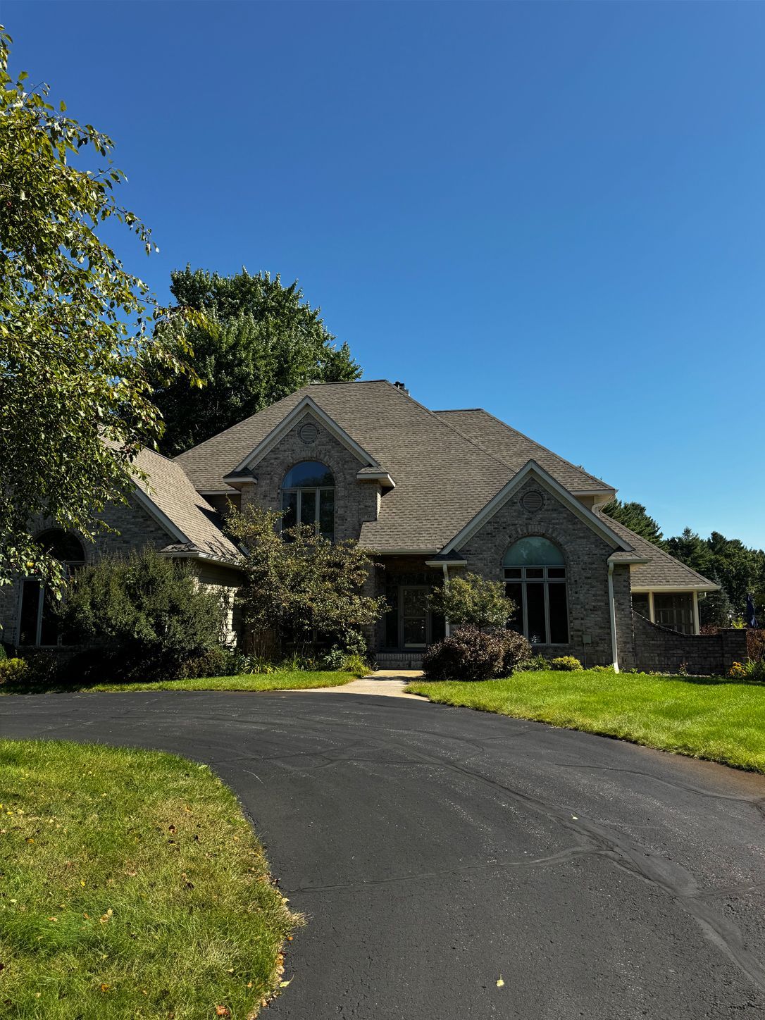 Stone house with a dark roof and arched windows, framed by trees and a curved driveway under a blue sky.