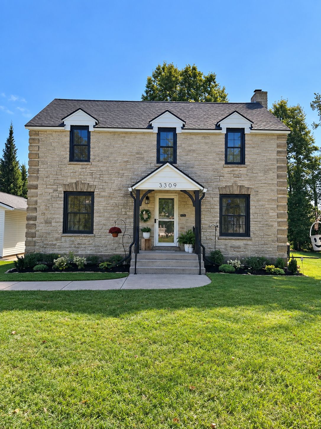 Two-story tan brick house with black-trimmed windows, a small front porch, and a green lawn under a blue sky
