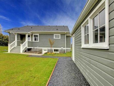 Gray house with white trim, front porch, and a gravel path beside a green lawn under a blue sky