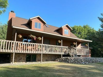 Log cabin house with a long front porch, stone foundation, and trees under a clear blue sky