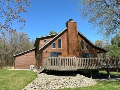 Rustic brown cabin with a wooden deck and stone foundation in a grassy yard under a blue sky
