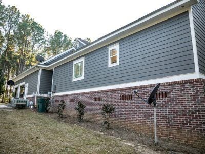 Gray house with red brick foundation and white-trimmed windows along a grassy side yard.