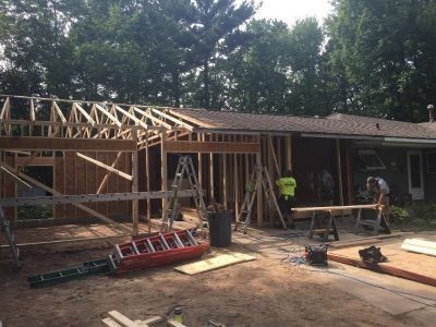House under construction with roof framing, ladders, and workers on a dirt lot.