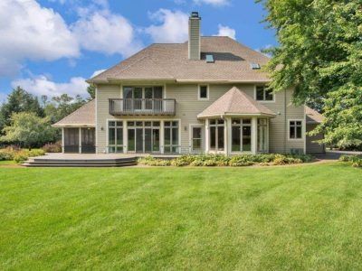 Large beige suburban house with a green lawn, deck, and trees under a blue sky