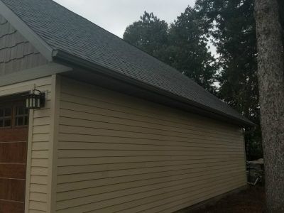 Side view of a beige house with a dark shingled roof and trees in the background