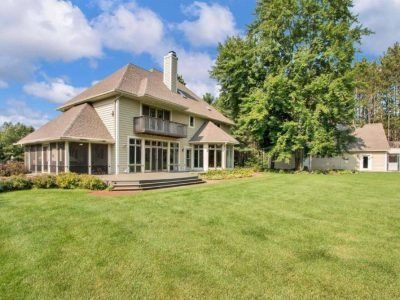 Large tan house with wraparound porch and chimney on a grassy lawn under a partly cloudy sky