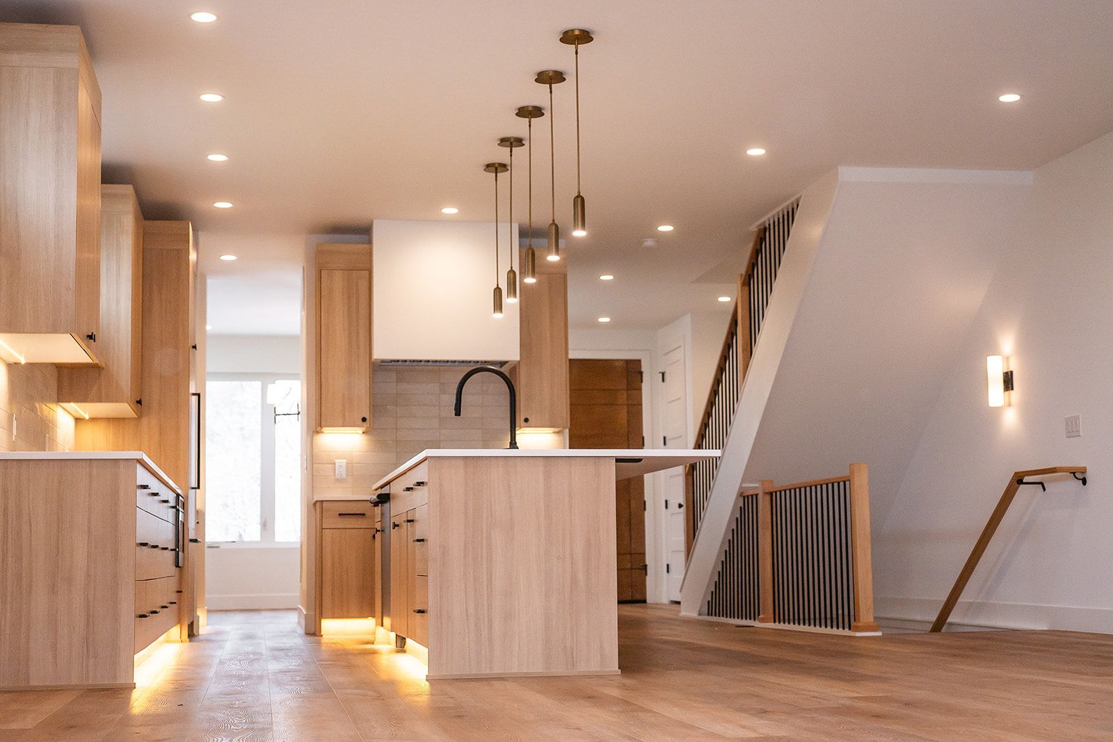 Modern kitchen with light wood cabinets, island, and pendant lights; staircase visible.