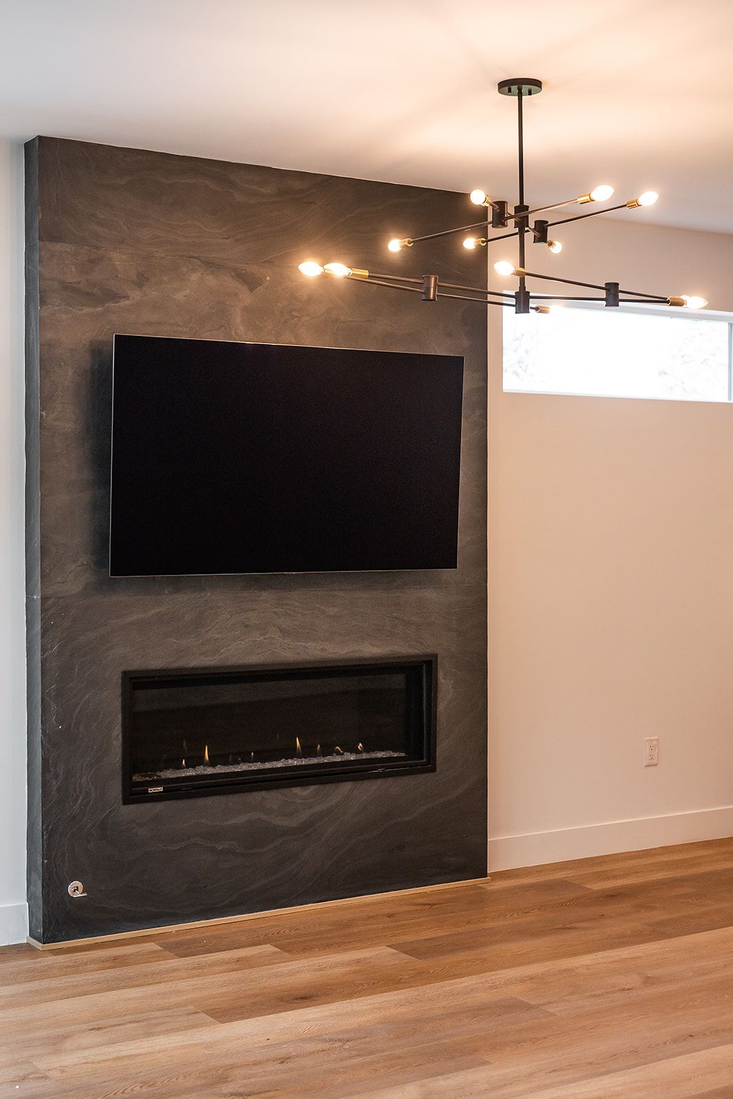 A modern fireplace and TV on a dark grey stone wall with a black light fixture and hardwood floor.