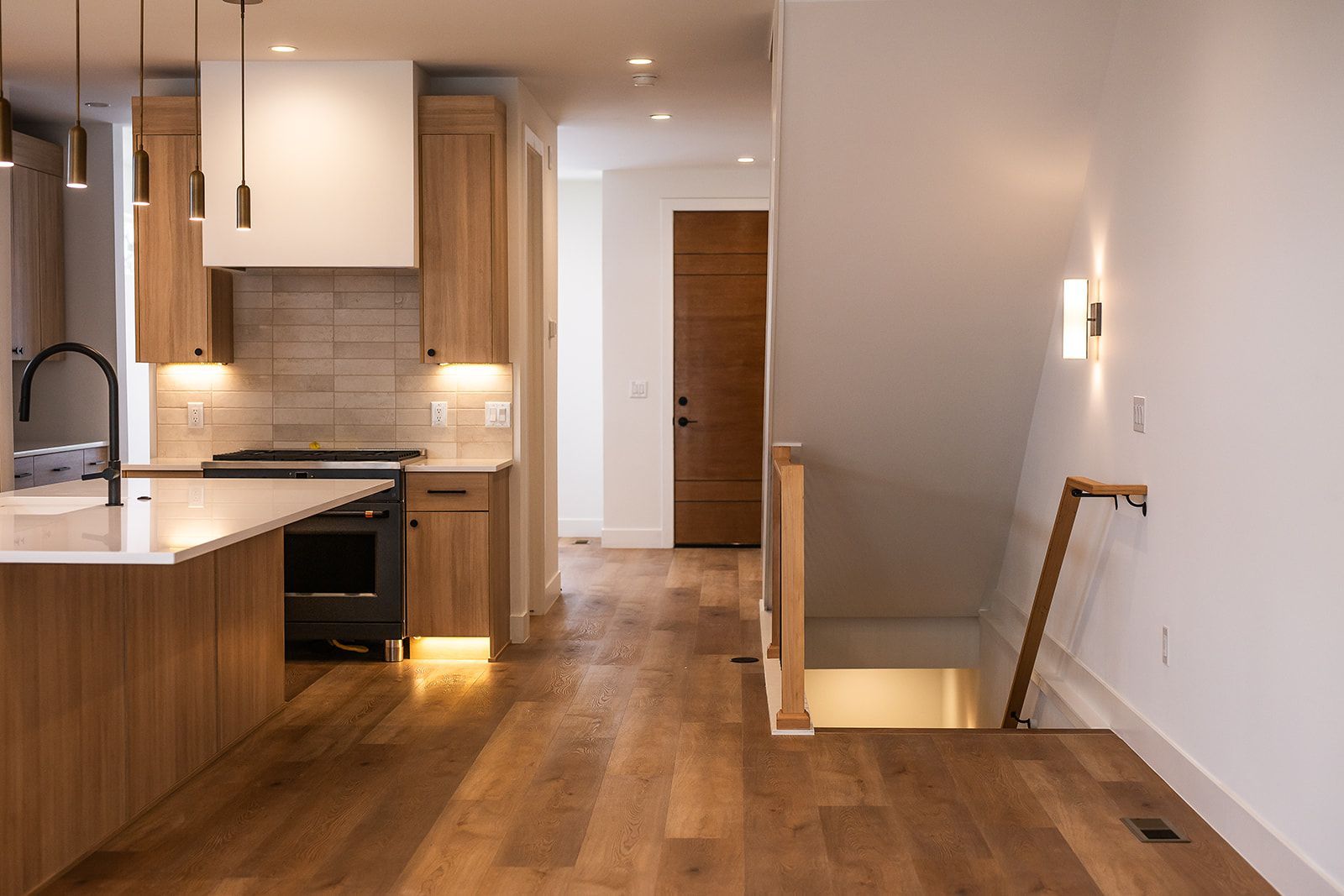 Modern kitchen with wood cabinets and flooring, stairs leading down, and a front door.
